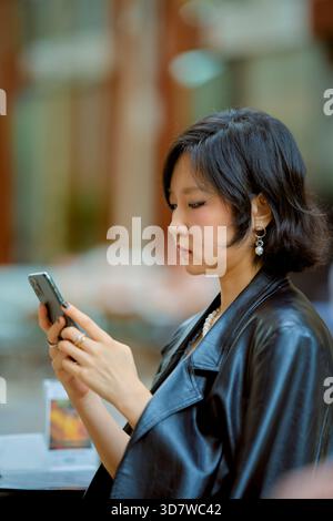 Eine junge Frau mit kurzen Haaren, die sich auf ihr Smartphone konzentriert und in einer städtischen Umgebung sitzt. Shanghai, China Stockfoto
