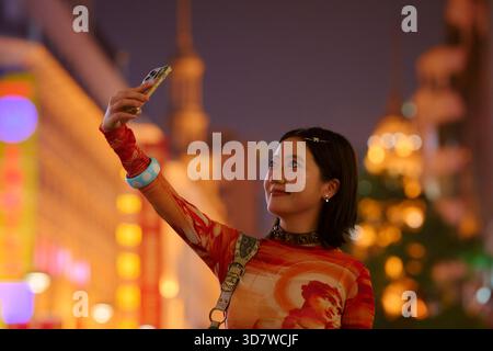 Eine junge Frau, die nachts ein Selfie vor einer lebhaften, bunten Kulisse der Stadt macht. Shanghai, China Stockfoto