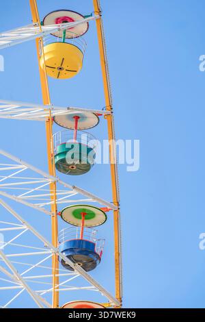 Detail von drei leeren Riesenradwagen (gelb, grün, weiß), die hoch oben an einem blassblauen Sommerhimmel hängen Stockfoto