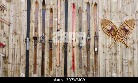 Eine Sammlung antiker Holzskier und traditioneller lederbezogener Schneeschuhe hängt senkrecht an einem rustikalen, alten Holzzaun. Dieses Gerät, Stockfoto