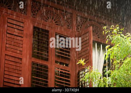 Eine Nahansicht des traditionellen malaiischen Holzfensters und der geschnitzten Lüftungsbleche bei Regenfällen. Das reiche rote Holz, die Lamellen und die Intri Stockfoto