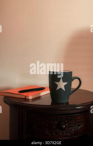 Grauer Becher mit orangefarbenem Notizbuch und einem Stift auf Holztisch. Stockfoto