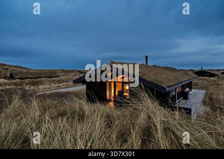 Rollende Gräser bedecken die Dünen, während einfache Holzhütten den Horizont säumen. Eine ruhige, offene Landschaft erinnert an Küstenzurückzug, Natur und ruhige Einsamkeit Stockfoto