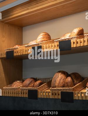Handwerkliche Brotpräsentation in der Bäckerei. Brotlaibe in Korbkörben auf Holzregalen in einer Bäckerei. Mehrere leere Etiketten sind für die Preisgestaltung sichtbar. Stockfoto