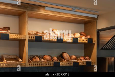 Handwerkliche Brotpräsentation in der modernen Bäckerei. Brotlaibe in Korbkörben auf Holzregalen in einer Bäckerei. Mehrere leere Etiketten für die Preisgestaltung. Stockfoto