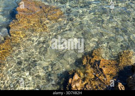 Blick von oben auf transparentes Meerwasser über Felsen; helles Tageslicht, schimmernde Reflexionen, gelb-braune Felsstrukturen, wellige Wasseroberfläche. Stockfoto