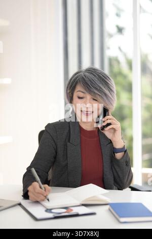 Asiatische ältere Erwachsene Geschäftsfrau verwaltet Aufgaben. Kommuniziert über das Mobiltelefon, schreibt effizient Notizen. Die professionelle Führungskraft übernimmt die Operationen von Stockfoto
