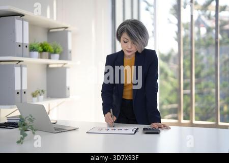 Reife asiatische Führungskraft steht auf der Analyse von Unternehmensleistungsdaten Finanzgrafiken mit Taschenrechner Stift Büro Arbeitsplatz Einstellung Geständnis Stockfoto