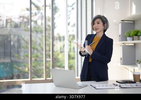 Nachdenkliche ältere asiatische Führungskraft steht im modernen Büro mit Notebook-Stift Planung Geschäftsprojekt erfolgreiches Karrierewachstum Innovationsstra Stockfoto