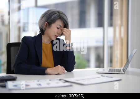 Reife asiatische Geschäftsführerin fühlt sich von den Anforderungen des Unternehmens überwältigt und gestresst, die fleißig an ihrem modernen Schreibtisch arbeiten Stockfoto