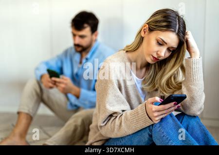 Ein junges Paar, das sich Rücken an Rücken lehnt, beide fokussiert auf ihre Smartphones und wirkt emotional distanziert Stockfoto