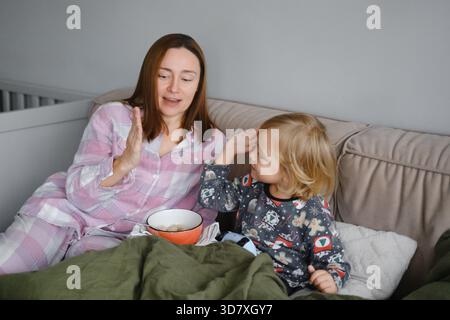 Mutter und Sohn frühstücken zusammen oder essen im Schlafanzug im Bett, Familienurlaub beim Frühstück im gemütlichen Schlafzimmer mit Kissen Stockfoto