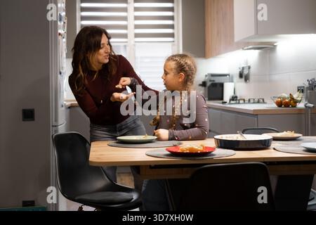 Mutter und Tochter essen gemeinsam hausgemachten Kuchen am Küchentisch, Nachtisch und Familienzusammenhalt in einem modernen Apartment Stockfoto