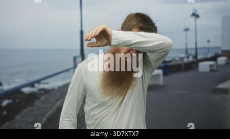 Bärtiger Mann in weißem Hemd wischt Schweiß von der Stirn auf der warmen sonnigen Strandpromenade am Meer ab; Erleichterung. Stockfoto
