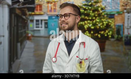Mann Doktor mit Stethoskop und Laborkitt lächelnd und auf einer nassen Straße in der Nähe einer dekorierten Ladenfront aufblickend; hoffentlich Sorgfalt Vertrauen. Stockfoto