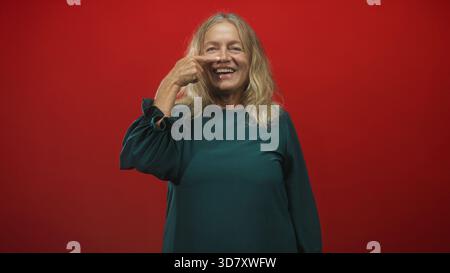 Die Seniorin zeigt Finger an Nase und lacht in grünem Kleid vor rotem Studio-Hintergrund; verspielte Freude. Stockfoto