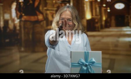 Die Seniorin zeigt mit dem Finger auf eine verpackte Geschenkbox mit blauem Band im prunkvollen Gebäude; Freude am Feiern. Stockfoto