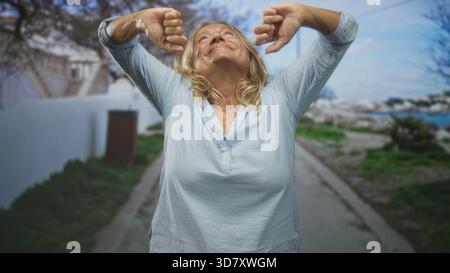 Ältere Frau mit ausgestreckten Armen auf einer Straße, Kopf nach hinten geneigt und Augen geschlossen, mit einer leichten Bluse; Zufriedenheit. Stockfoto