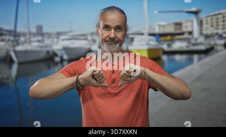 Ein Mann mit langen grauen Haaren und Bart zeigt mit beiden Händen die Daumen nach unten an der Marina Street an Booten und am Pier in der Nähe des Hafengehäuses; Missbilligung. Stockfoto