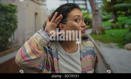 Frau, die Hand an Ohr schröpft, indem sie auf dem Parkweg baut, Brille und gemusterte Jacke trägt; Neugier hört aufmerksam zu. Stockfoto