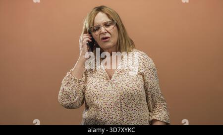 Frau mittleren Alters, die das Smartphone am Ohr hält, die Hand am Telefon hochgehoben, mit offenem Mund und einer Brille vor einem nahtlosen Studio-Hintergrund grimmend; f Stockfoto