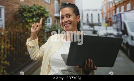 Frau, die den Laptop hält, zeigt mit dem Finger nach oben auf der Straße mit Bürgersteig und geparktem Van am Ziegelgebäude; Zuversicht Optimismus. Stockfoto