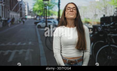 Frau mit Brille lächelt mit geschlossenen Augen und Händen locker an der Taille auf einer Straße neben geparkten Fahrrädern und Autos; ruhige Zufriedenheit. Stockfoto