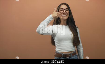 Frau mit langen Haaren und Brille zeigt zwei Daumen nach oben, zeigt sichtbare Hände und ein breites Lächeln, trägt ein weißes Hemd, Jeans und Gürtel in der Studiowand Stockfoto