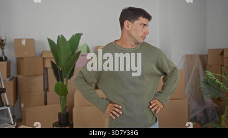 Junger Mann steht mit Händen auf Hüften neben gestapelten Pappschachteln im Innenraum des Gebäudes und trägt einen olivfarbenen Pullover; Unsicherheit. Stockfoto