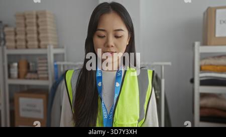 Eine junge Frau in einer reflektierenden Weste macht eine Herzgeste in einem Freiwilligenzentrum, umgeben von Hilfsgütern, die Gemeinschaft und Freundlichkeit betont. Stockfoto