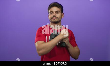 Junger Rettungsschwimmer, der ein Fernglas und eine Pfeife hält, rotes Hemd trägt, im Studio mit violettem Hintergrund steht; Wachdienst. Stockfoto