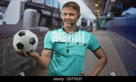 Schiedsrichter hält Fußball in der Hand, Pfeife sichtbar und Headset-Mikrofon im Gebäudekorridor; Vertrauensmacht. Stockfoto