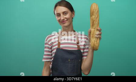 Frau mit Schürze und Einweghandschuhen, die ein Baguette hält und in einem türkisfarbenen Studio lächelt; freundliche Gastfreundschaft. Stockfoto
