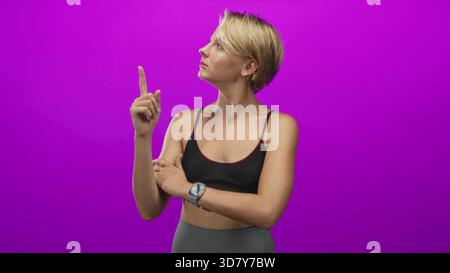 Frau zeigt den Zeigefinger nach oben mit sichtbarer Smartwatch und Sport-BH im Studio; selbstbewusste Haltung. Stockfoto
