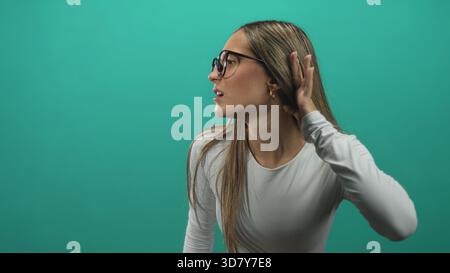Frau mit Brille, die Hand an Ohr schröpft und den Kopf leicht dreht im grünen Studio; Neugier Hörfokus Konzentration. Stockfoto