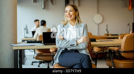 Eine selbstbewusste Frau in einem grauen Anzug genießt einen Moment der Freizeit in einem modernen Büro. Sie lächelt im Sitzen, umgeben von Leuten, die im bac zusammenarbeiten Stockfoto