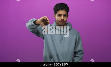 Mann in grauem Hoodie zeigt Daumen nach unten mit sichtbarem Daumen und neutralem Ausdruck im Studio; Ablehnung der Ablehnung. Stockfoto