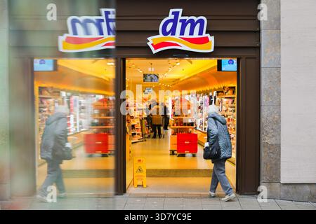 Bamberg, Deutschland 27. November 2025: Im Bild: Blick in den Eingangsbereich einer dm Drogerie in der Bamberger Innenstadt. Vor dem Geschäft kommt ein Fahrzeug oder Objekt vorbei und erzeugt Bewegungsunschärfen im Vordergrund, während sich die Kunden im Inneren befinden Stockfoto