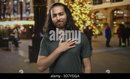 Junger Mann mit rechter Hand auf der Brust auf der Straße vor dem beleuchteten Weihnachtsbaum und verschwommenen Einkäufern, kurze Halskette sichtbar, graues T-Shirt trägt; Dankbarkeit. Stockfoto