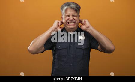 Polizist stopft Ohren mit Fingern, grimmig und zuwinkelt in einem orangen Studio mit sichtbarem Schild und Funkkabel; Unannehmlichkeit Ärger. Stockfoto