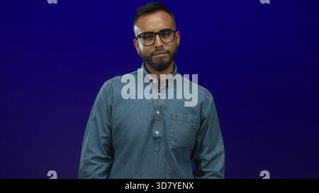 Junger hispanischer Mann, der Denim-Hemd mit Brille trägt, beugt den Arm und zeigt Bizeps im blauen Studio; Selbstvertrauen. Stockfoto