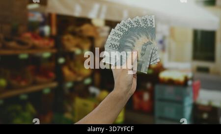 Kaukasischer Mann, der indonesische Rupiah-Banknoten in einem Supermarkt hält, mit Obst im Hintergrund als Symbol für Shopping und Geldwechsel. Stockfoto