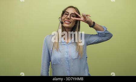 Junge Frau Callcenter-Vermittler mit Headset blinkendes Friedensschild und lächelndes Lächeln im Studio vor hellgrünem Hintergrund; spielerisches Selbstvertrauen. Stockfoto