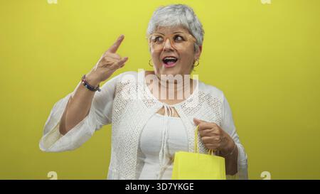 Frau mit grauen Haaren und Brille, die gelbe Einkaufstasche mit dem Zeigefinger nach oben hält, in weißer Bluse im Studio; fröhliches Selbstvertrauen. Stockfoto