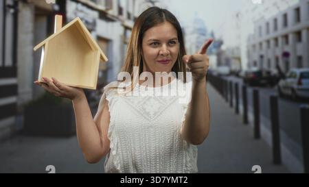 Eine Frau, die ein Holzmodell auf der Straße hält, zeigt den Finger nach vorne, während sie lächelt; Selbstvertrauen beim Hausbesitz. Stockfoto