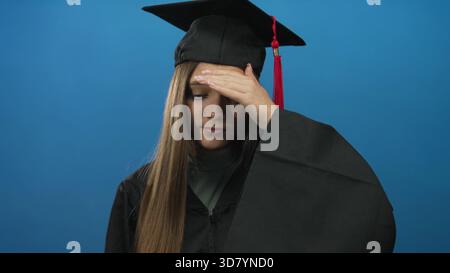 Teenager-Mädchen im Mortarboard- und Abschlusskleid hält Hand auf der Stirn im blauen Studio; Unsicherheit Zweifel reflektieren Zögern. Stockfoto