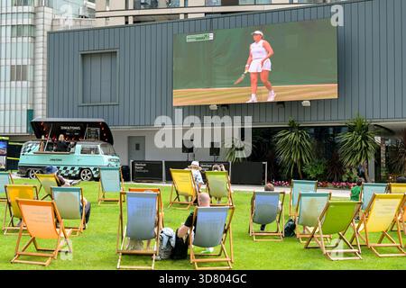 London, England, Großbritannien - 4. Juli 2025: Tennisfans sitzen auf Liegestühlen auf einem Rasenplatz in Paddington und beobachten auf einer Großbildleinwand Tennis in Wimbledon. Stockfoto