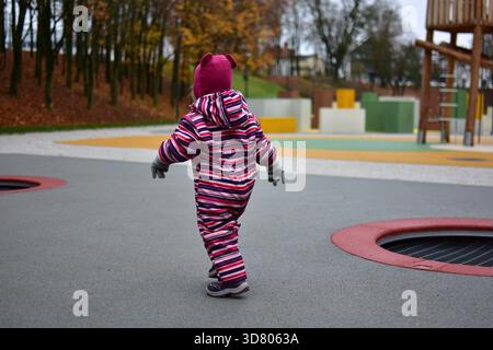 Kleinkind Mädchen in einem gestreiften Winter insgesamt gehen selbstbewusst über einen modernen Outdoor-Spielplatz mit hellem Gummiboden und Holzspielstrukturen Stockfoto