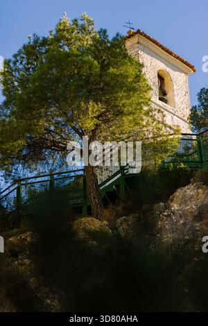 Grüner Baum im Fokus vor einer ruhigen Kirche und Glockenturm unter einem hellblauen Himmel an einem sonnigen klaren Tag, vertikale Landschaft. Stockfoto