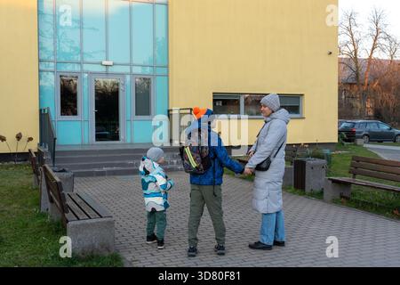 Mutter mit zwei Söhnen, die zum Eingang des Schulgebäudes gehen, Hände an kalten Herbstmorgen in Winterjacken halten. Das Konzept des Familienschulalltags Stockfoto